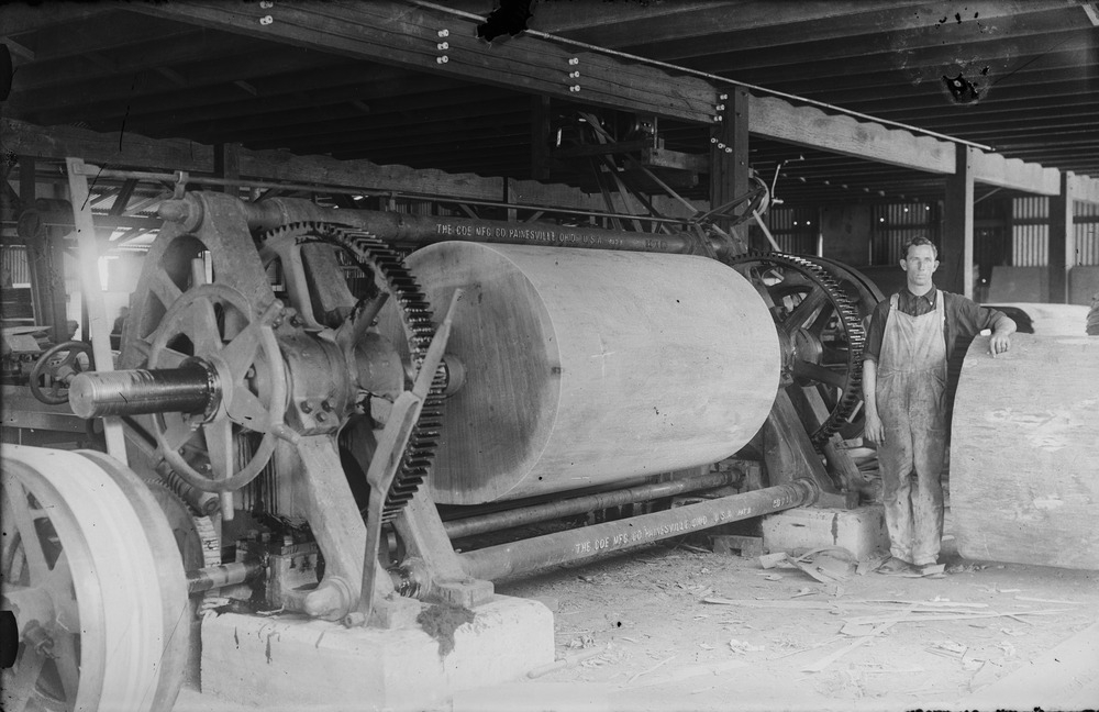 A 1935 glass plate photograph of a huge log mounted on a wood lathe inside a timber mill