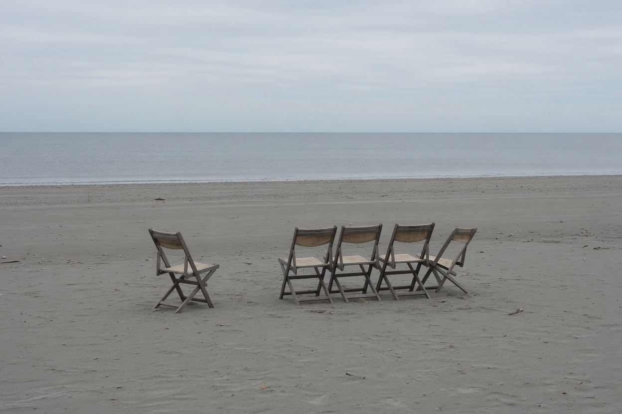 Five plain wooden folding chairs on grey sand facing a flat grey sea under an overcast sky, one chair set a little apart, nobody on the beach.