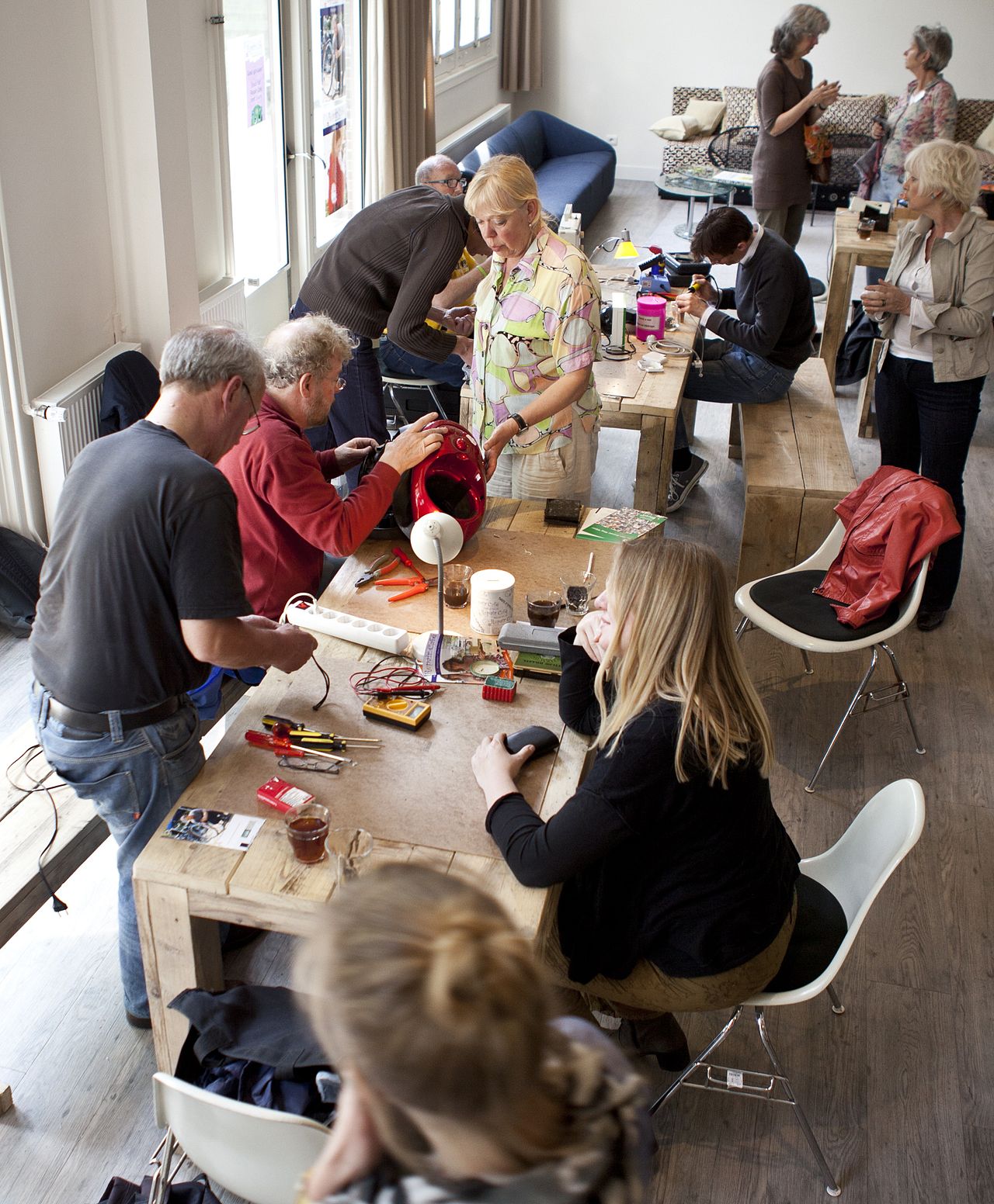 A busy repair morning: volunteers working together at long wooden tables, fixing a red vacuum cleaner and small appliances, with tools and cups of tea spread across the tables.