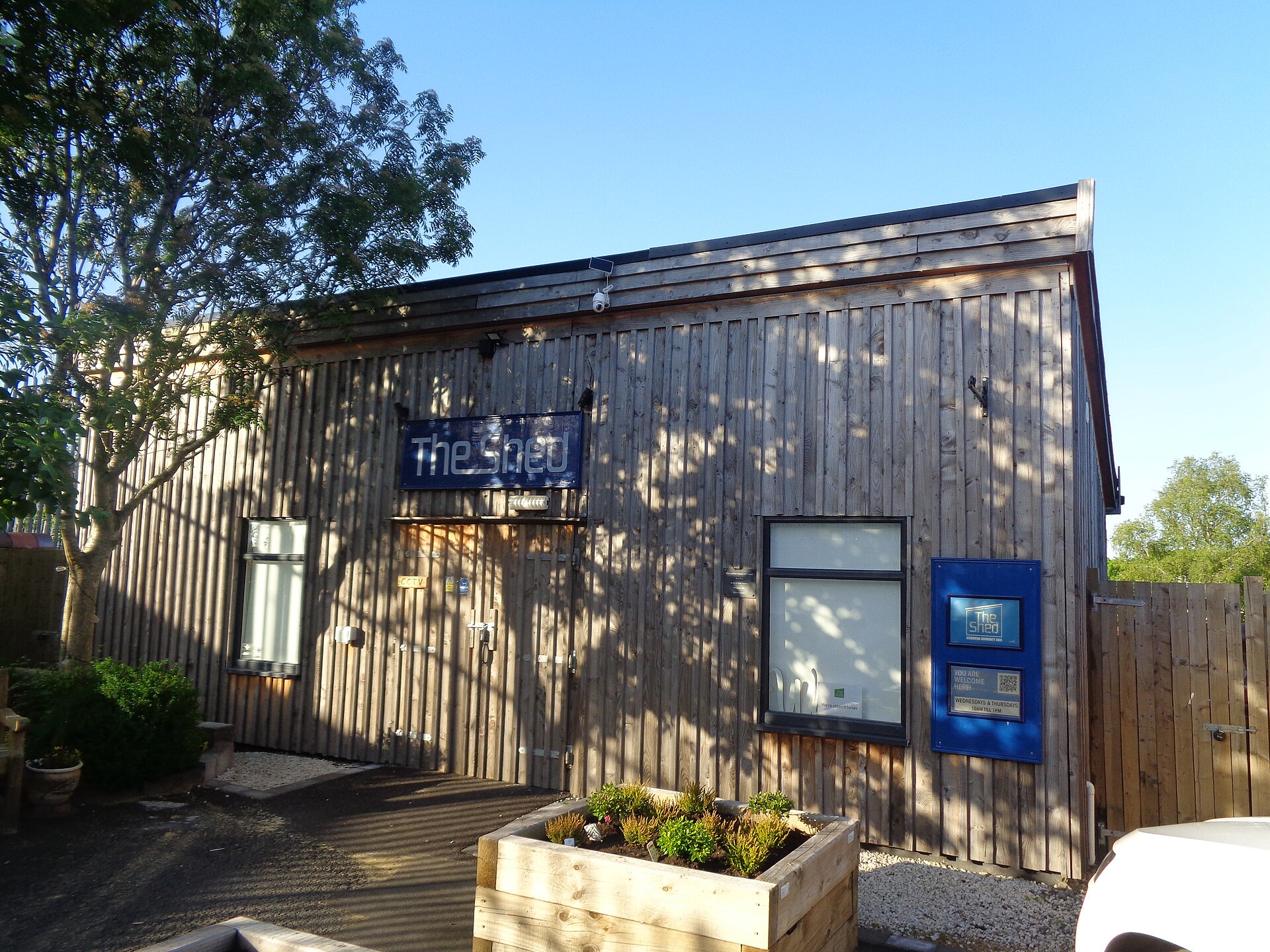 A timber-clad community shed on a sunny day, with a blue sign reading The Shed above the door, a noticeboard by the window, and a planter box out the front.