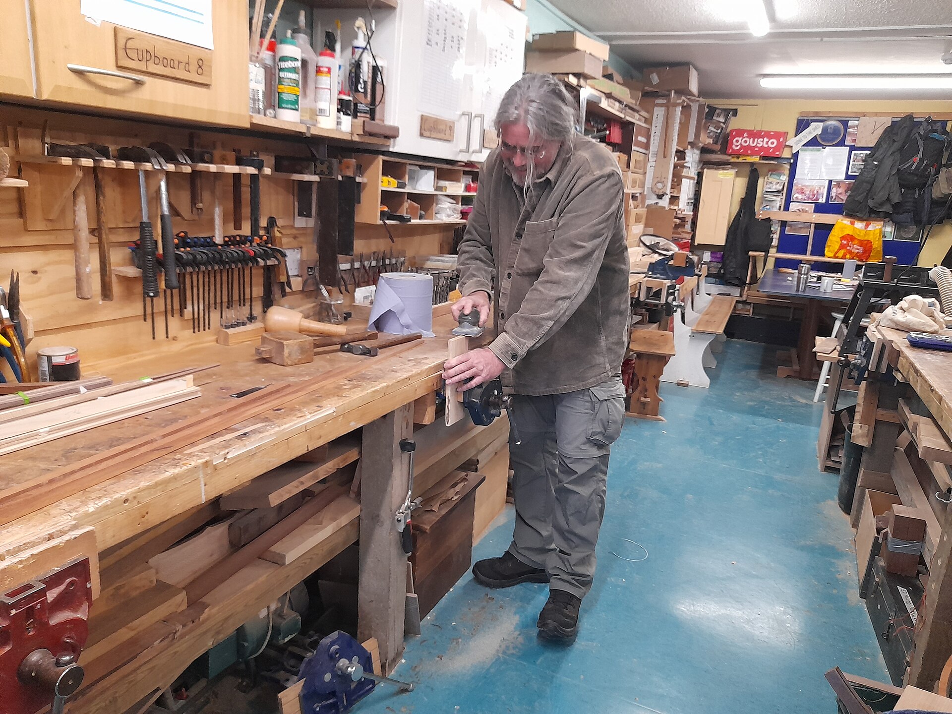 A man with long grey hair planing a piece of timber held in a bench vice, at a long crowded workbench with clamps, mallets and hand tools hung on the wall behind him.