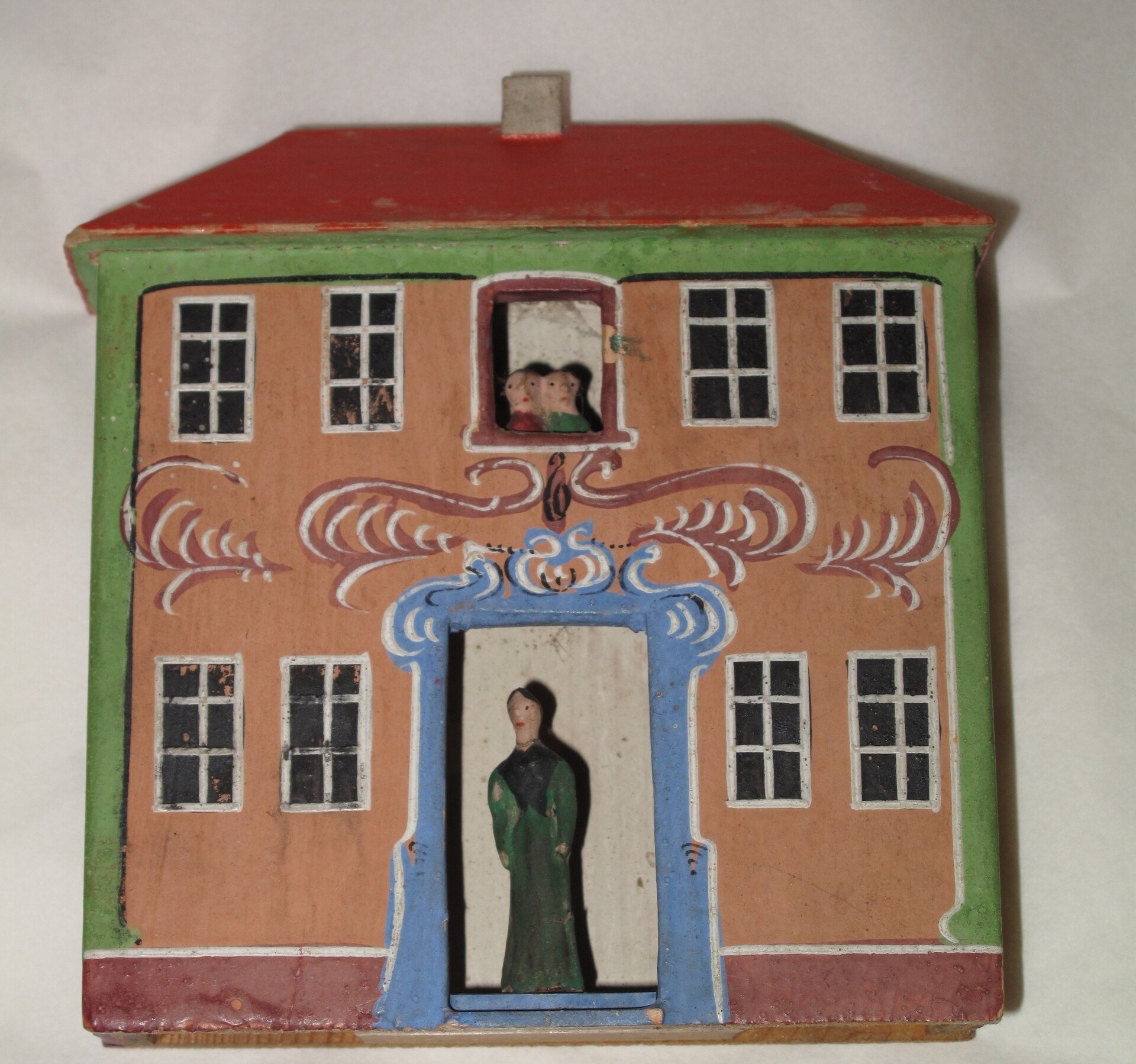 An old painted wooden toy house with a red roof, decorative painted flourishes, and small carved figures in the doorway and upstairs window.